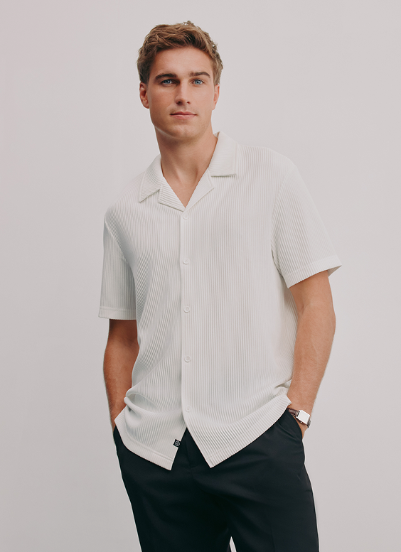A young man with light brown hair and blue eyes standing with hands in pockets against a plain light gray background. He is wearing a white, short-sleeved, pinstripe button-up shirt and black pants.