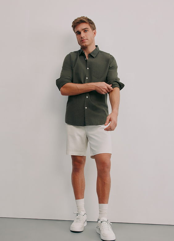 Young man with casual style, wearing a dark green button-up shirt, white shorts, white sneakers, and white socks, standing against a plain white background.