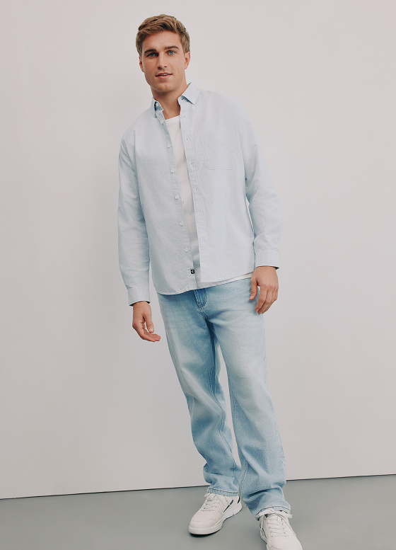 Young man standing against a plain white background, wearing a light blue button-down shirt, light wash jeans, and white sneakers.