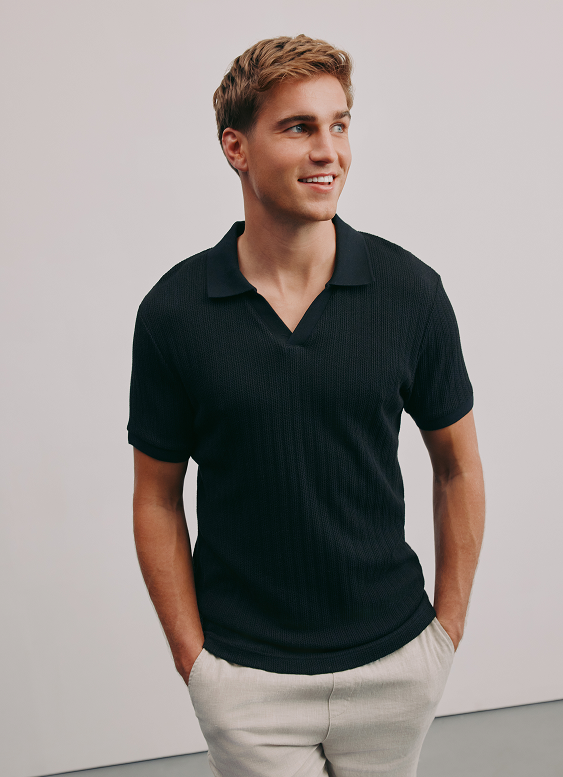 A young man with light brown hair, dressed in a black polo shirt and light-colored pants, standing against a plain light gray background, smiling and looking to his right.