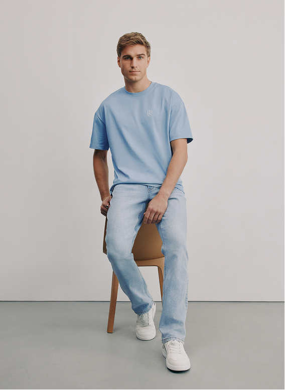 A young man in a light blue T-shirt and jeans sitting on a wooden chair against a plain gray background.