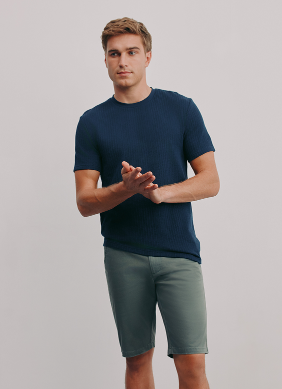 Young man in a navy blue t-shirt and green shorts standing against a plain gray background.