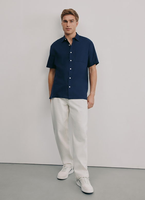Young man standing against a plain light gray wall, wearing a navy blue short-sleeve button-up shirt, white pants, and white sneakers.