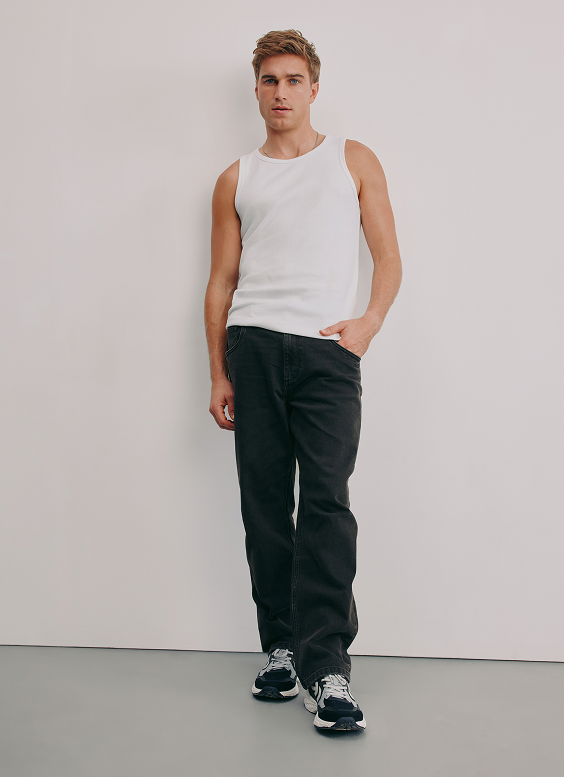Young man wearing a white sleeveless tank top, black jeans, and running shoes, standing against a plain white background.