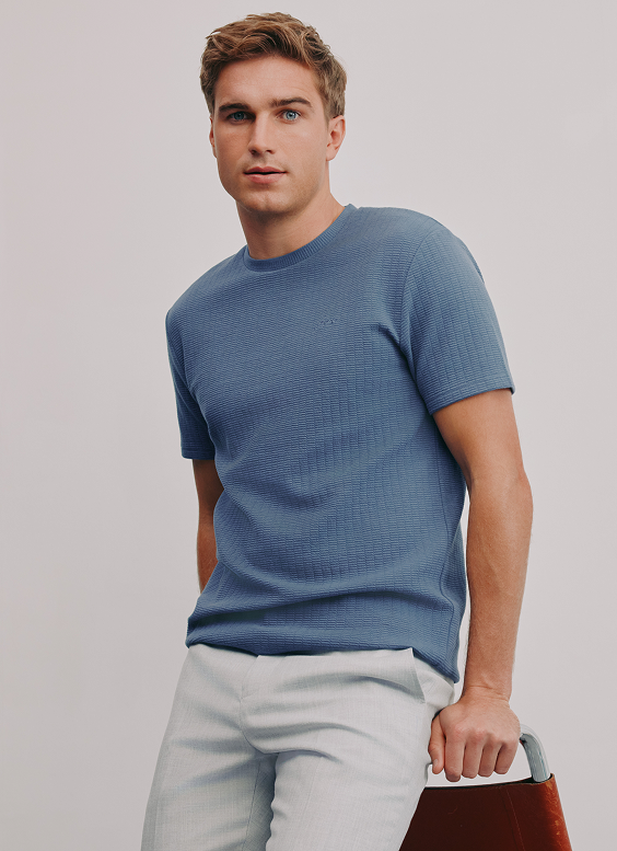 A young man with light skin, blue eyes, and short wavy brown hair posing against a plain gray background. He is wearing a blue textured short-sleeve shirt and light-colored trousers, resting his right hand on the back of a chair.