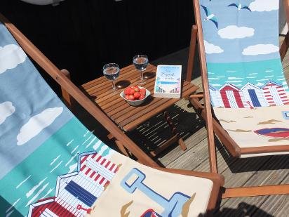 Beach-themed patio setup with wooden lounge chairs with striped cushions, a small table with two glasses of wine, strawberries, and a card, all against a backdrop of beach artwork and a black fence.