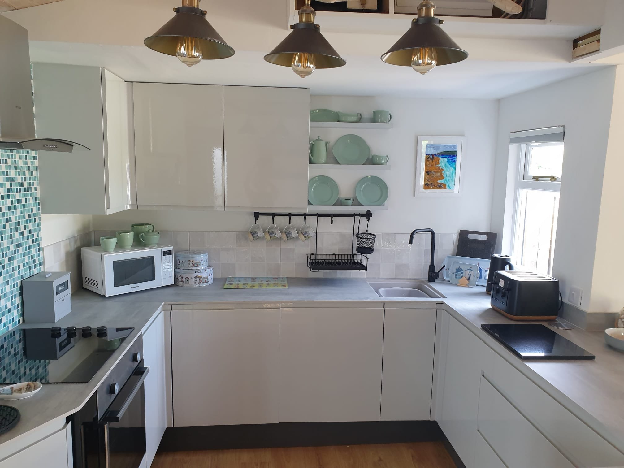 Kitchen with white cabinets, open shelving with mint green dishes, black pendant lights, microwave, toaster, stove, and small window.
