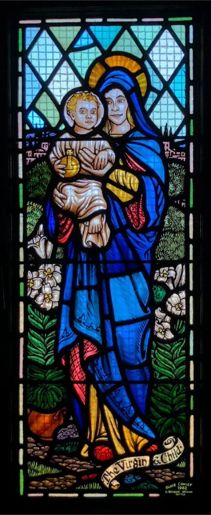 Colorful stained glass window depicting the Virgin Mary holding baby Jesus, surrounded by flowers and greenery, with a banner reading 'The Virgin & Child' at the bottom.