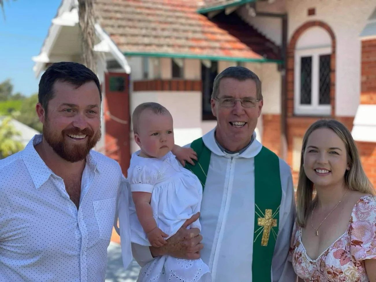 Group of four people, two men, a woman, and a baby, standing outdoors in front of a house with a newsboy cap and smiling.