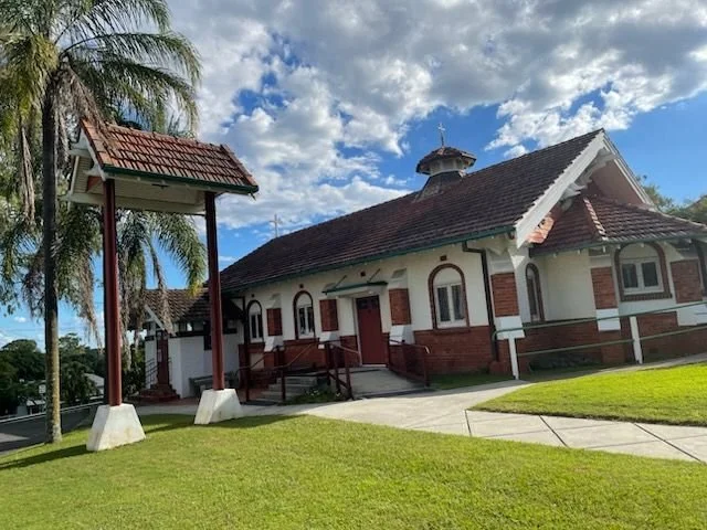 Church building with white walls, red brick accents, a brown tiled roof, and a small steeple, surrounded by a well-maintained lawn, palm trees, and a cloudy blue sky.