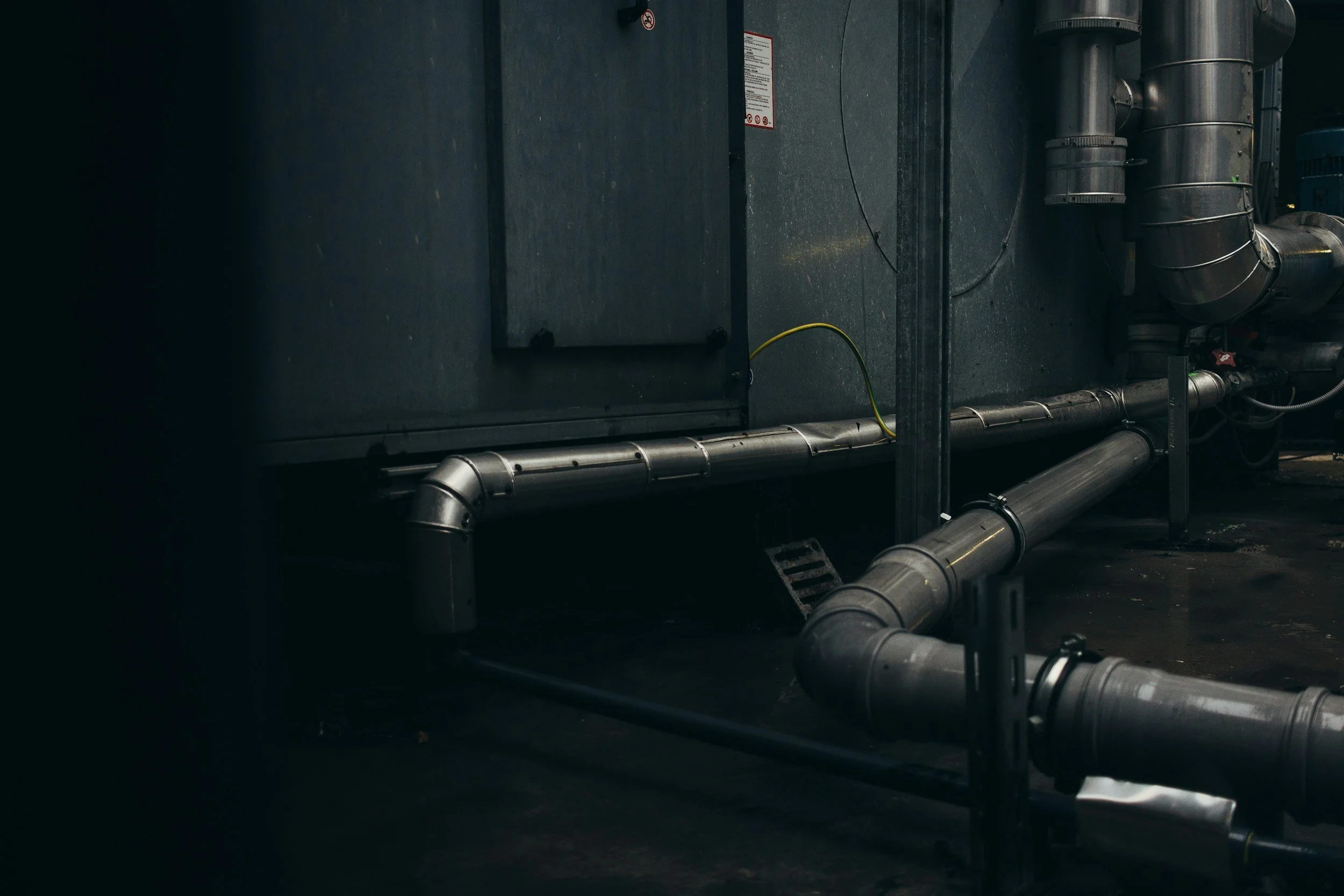 Industrial setting with large black pipes, a metal electrical panel, and a yellow wire connected to a dark wall.