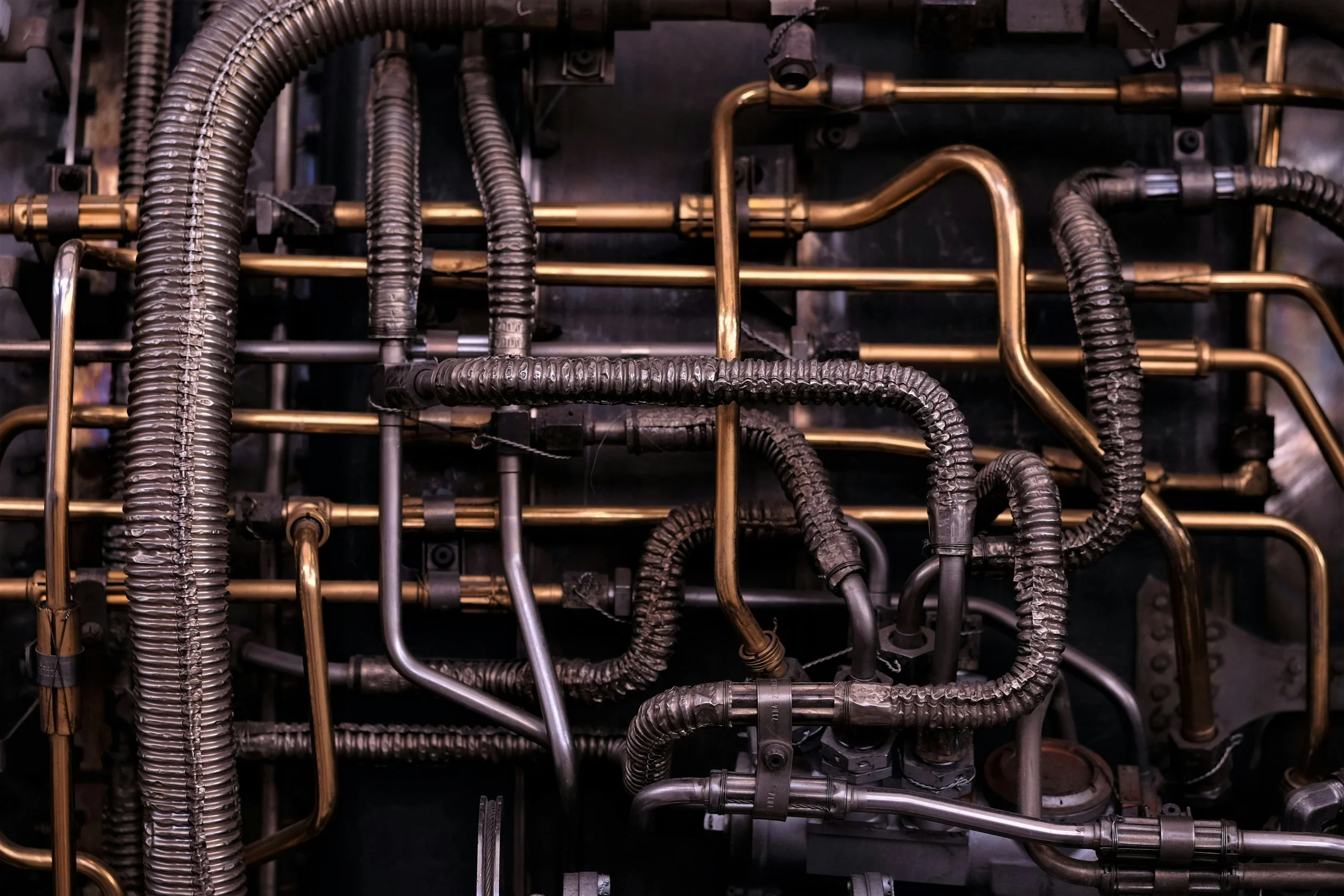 A close-up view of a complex network of metallic pipes and hoses, with some coated in a black protective layer and others in a brass or silver finish, forming part of machinery or industrial equipment.
