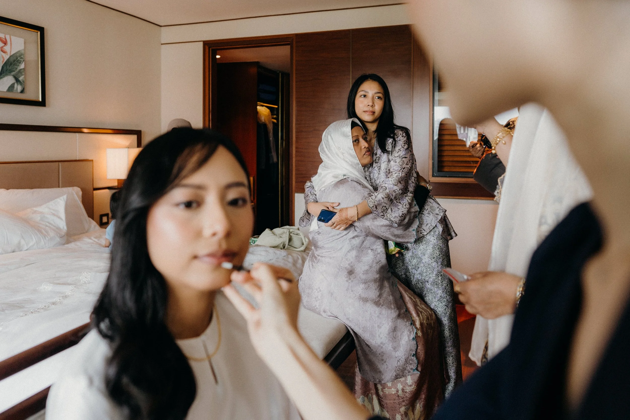 A group of women in a hotel room, with one woman applying lipstick, another sitting on the bed, and two others embracing near a closet, all dressed in traditional clothing.