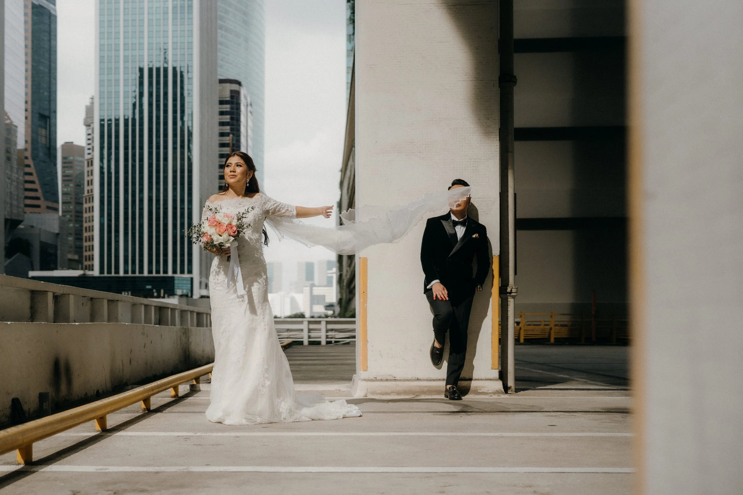 Bride in a white wedding dress holding a bouquet, extending her arm as a man in a black tuxedo, blindfolded, leans against a wall in an urban parking garage with Singapore's urban backdrop.