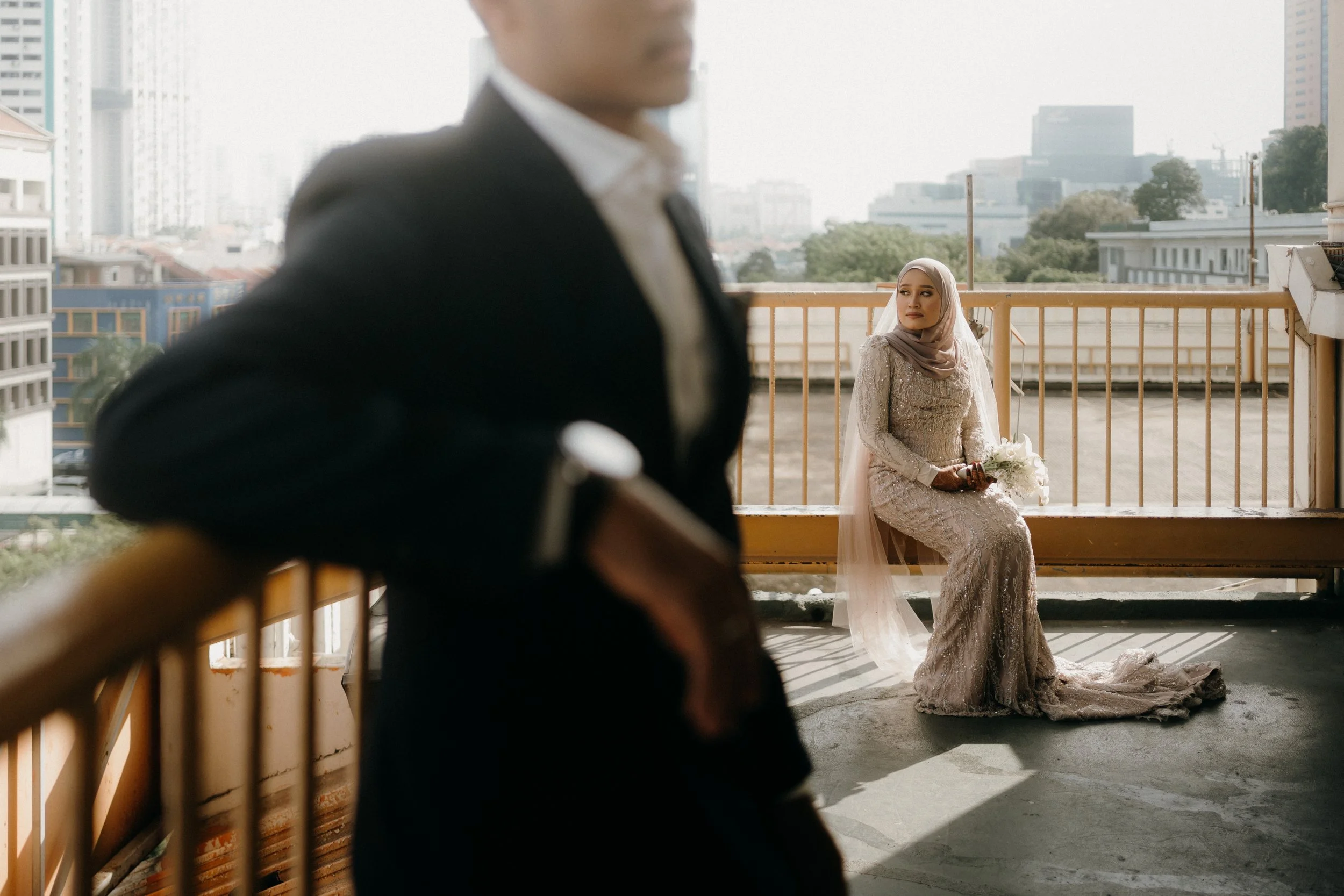 A woman in a wedding dress sitting on a bench on a balcony, holding a bouquet, looking contemplative, with a man in a suit standing in the foreground, blurred.