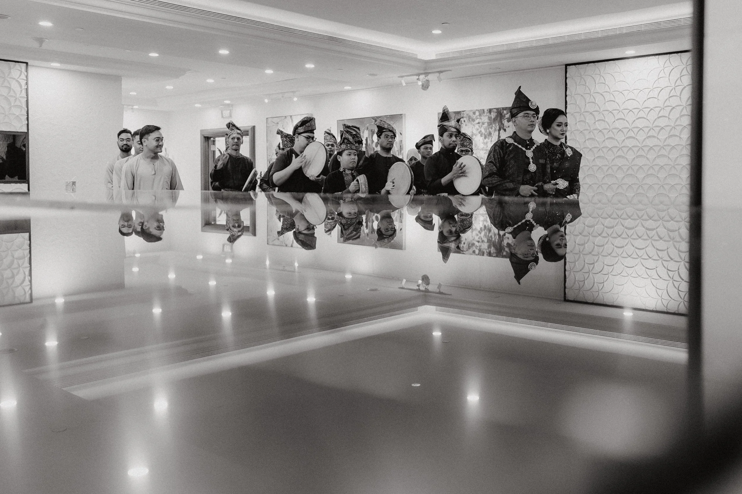 A group of people dressed in traditional attire, some playing drums, standing indoors with a reflective surface in the foreground.
