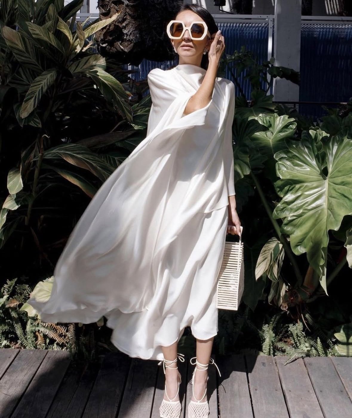 Woman in a flowing white dress with large white sunglasses, standing outdoors on a wooden surface surrounded by large green tropical leaves, holding a woven clutch and wearing high-heeled shoes.