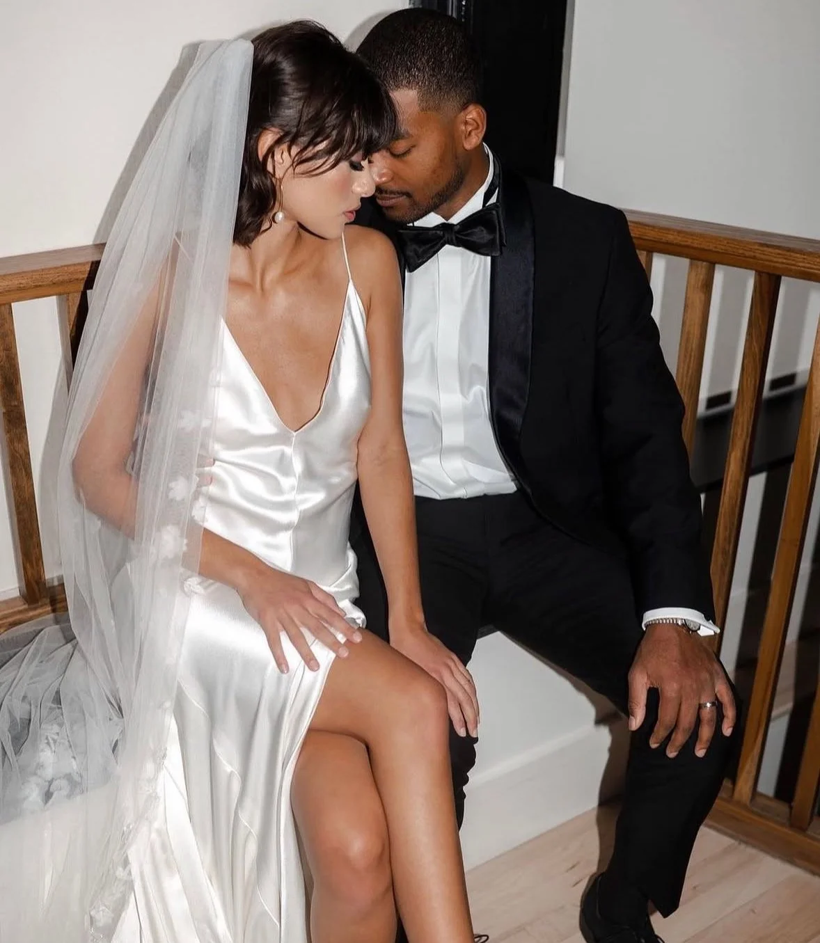 A bride and groom sitting close together, with heads touching, dressed in wedding attire. The bride is wearing a satin wedding dress and a veil, and the groom is in a black tuxedo with a bow tie.