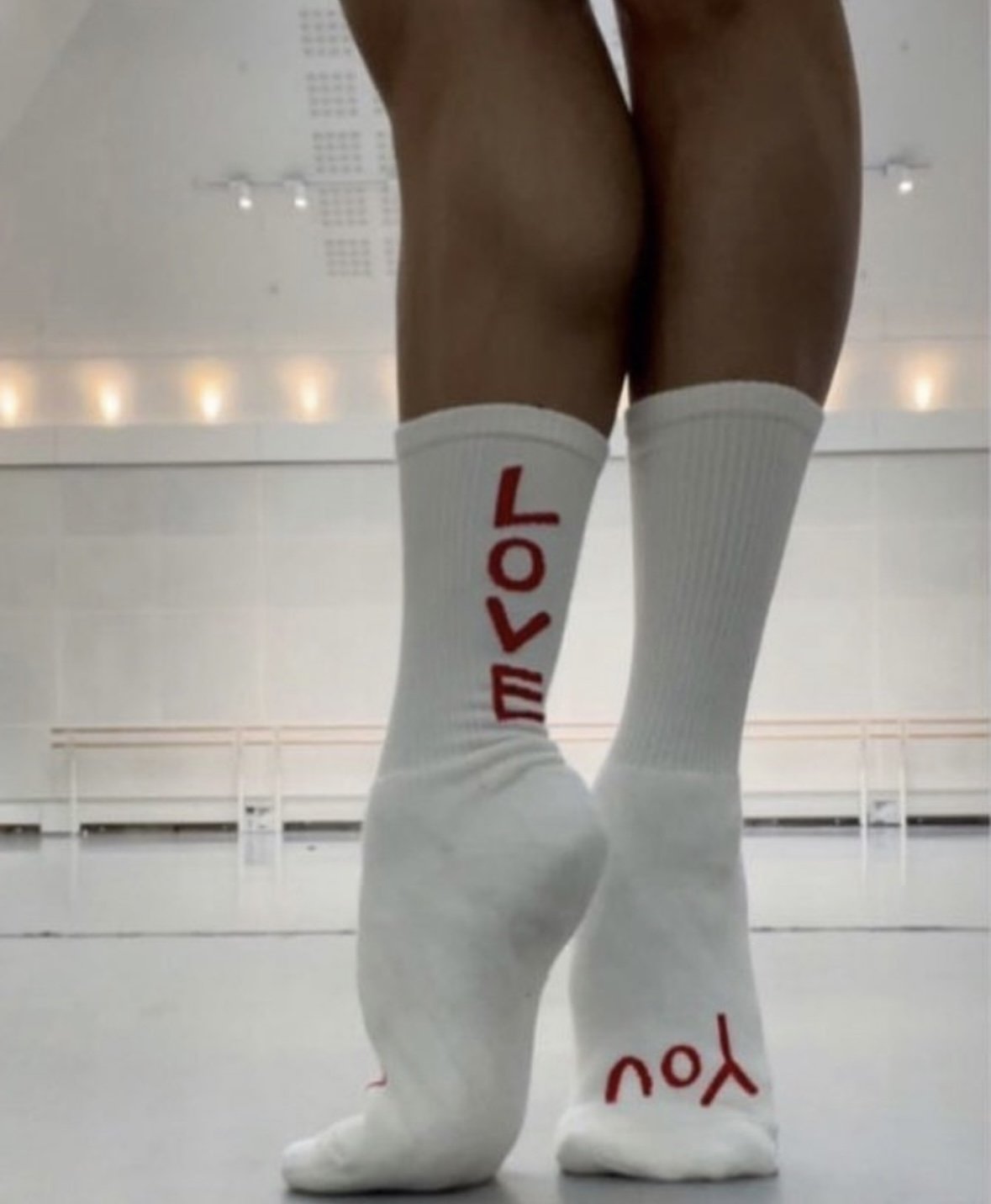 A person wearing white knee-high socks with red words that say "YOU" and "LOV" on each sock, viewed from behind in a dance or sports studio.