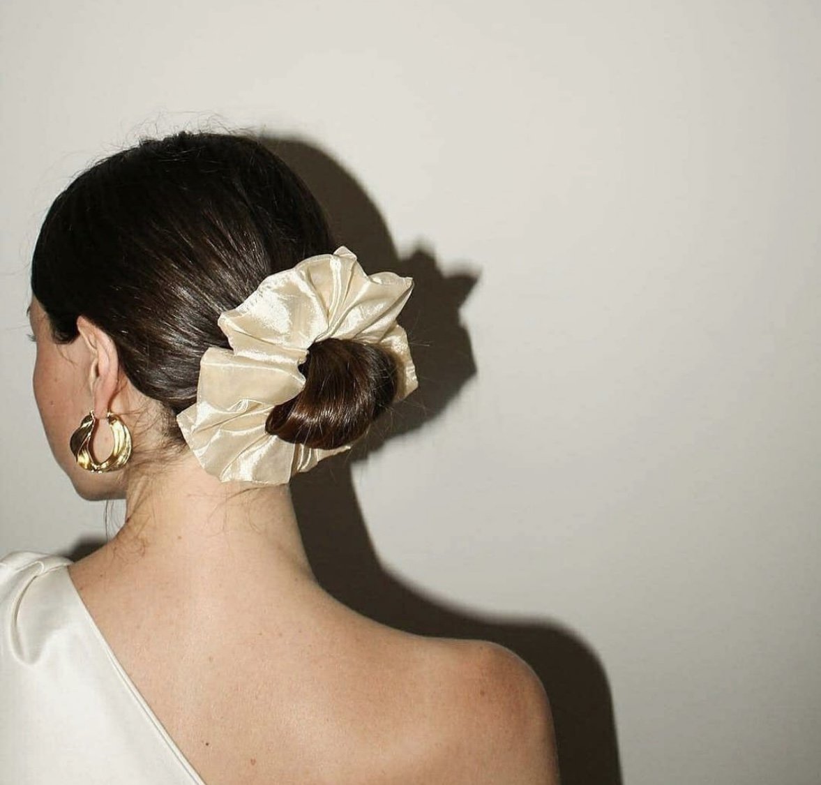 A woman with dark brown hair styled in a bun decorated with a large, beige, silky scrunchie. She is wearing gold hoop earrings and a white top, with her back turned to the camera against a plain, light-colored wall.