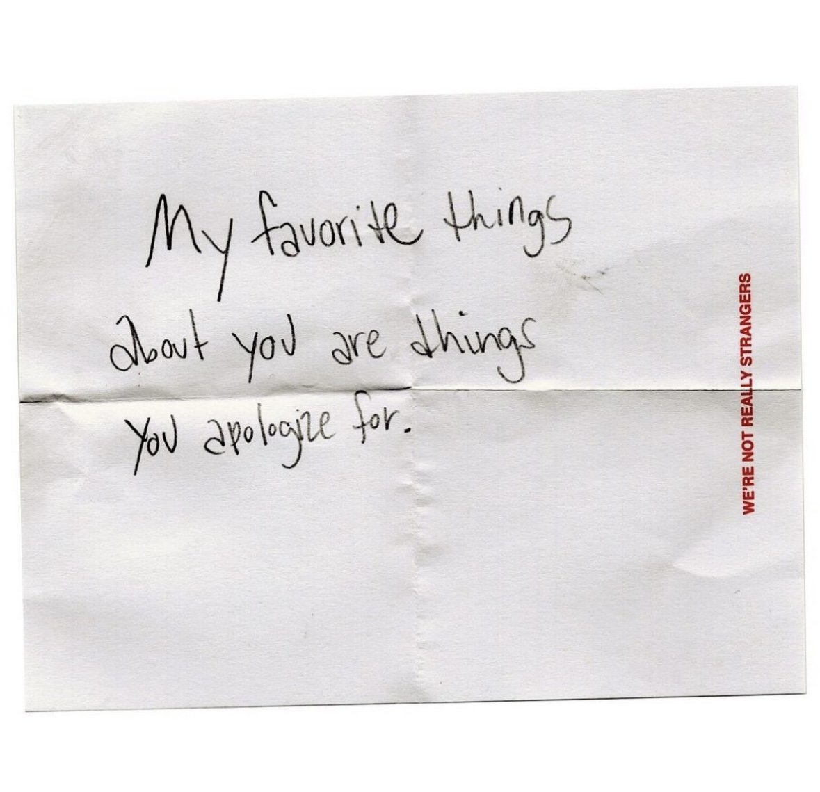 A handwritten note on a white piece of paper that says, 'My favorite things about you are things you apologize for.' The paper has printed red text along the right edge that reads, 'WE'RE NOT REALLY STRANGERS'.