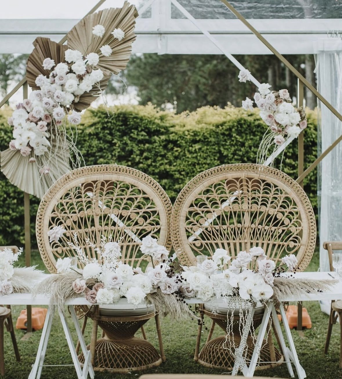 Decorated outdoor wedding table with two rattan chairs, floral arrangements of white and blush flowers, and a geometric floral backdrop with draped fabric and greenery.