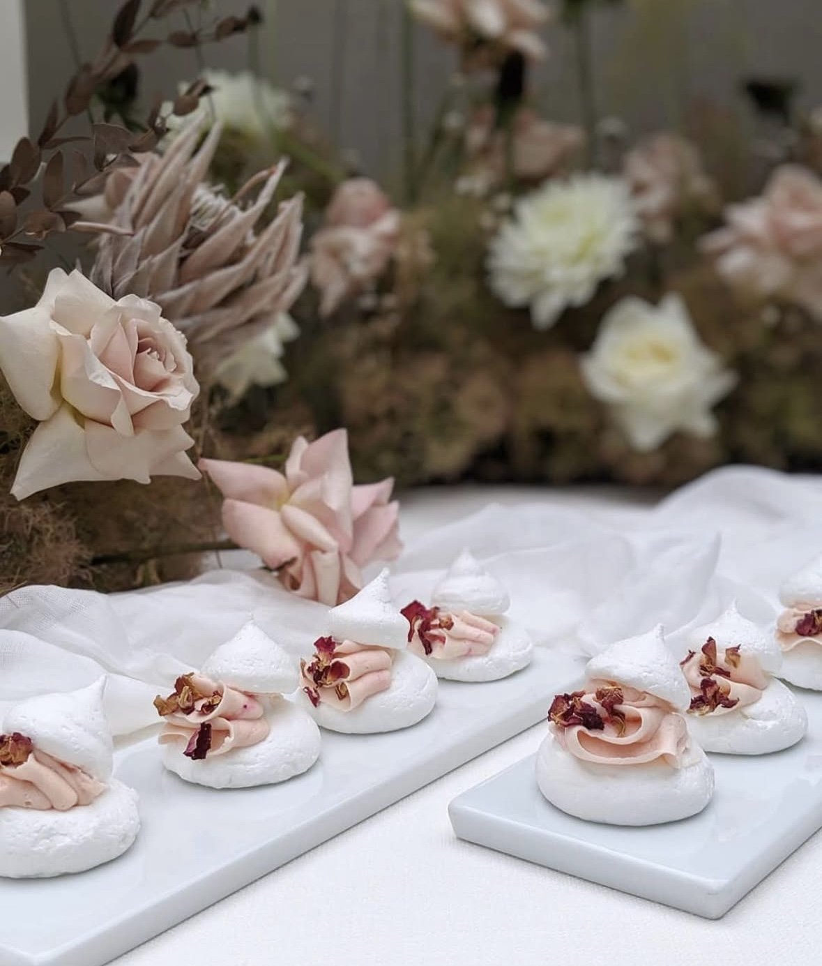 Decorative white meringue cookies topped with pink cream and dried rose petals, arranged on white platters, with pastel pink and white flowers in the background.