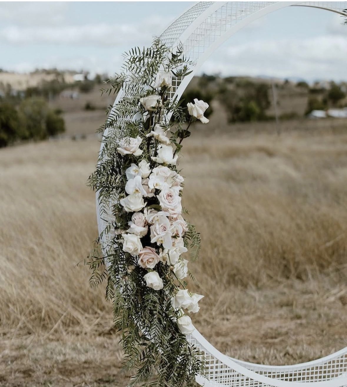 Close-up of a decorative floral arrangement with white and pale pink roses and greenery attached to a circular metal arch outdoors, with a rural landscape in the background.