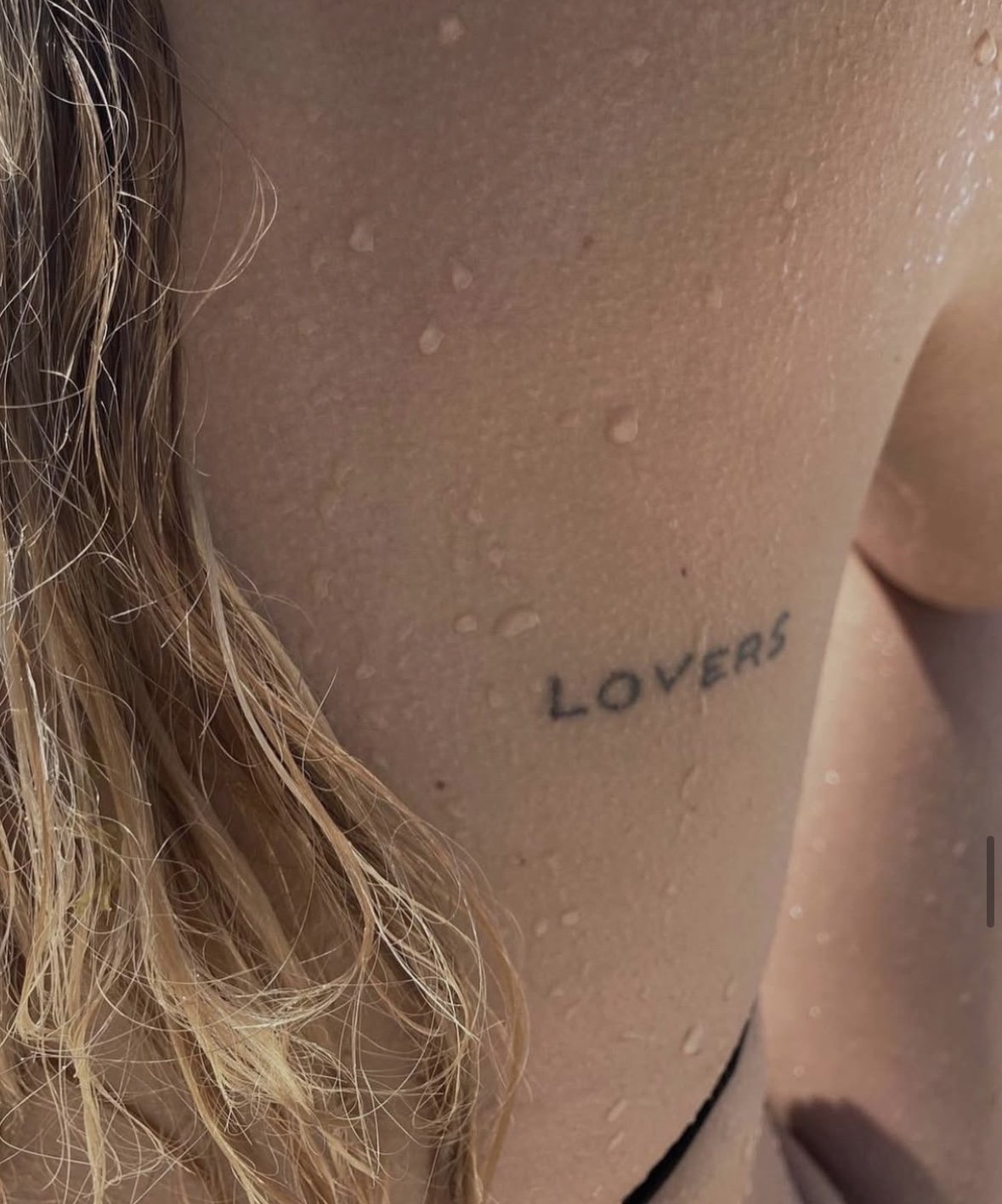 Close-up of a person's shoulder with water droplets and a tattoo that says 'LOVERS' near the collarbone.