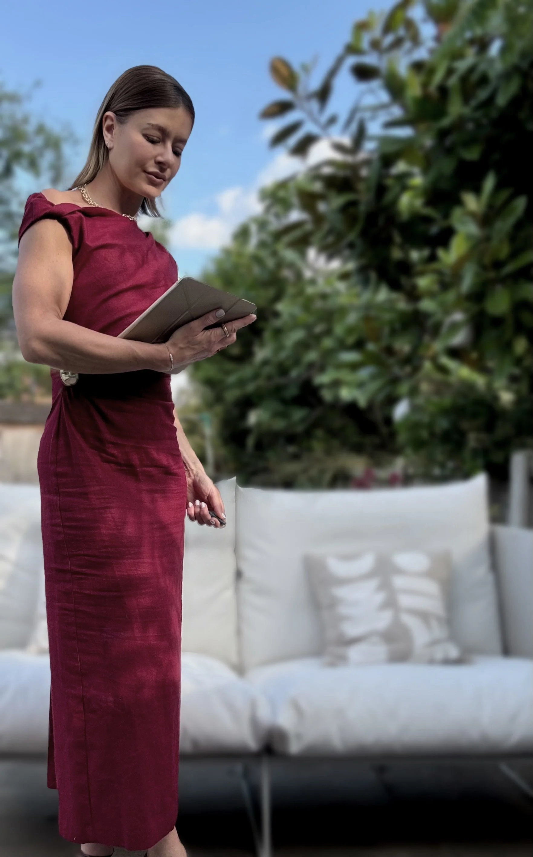 A woman in a red dress standing outdoors on a patio with white cushions, holding a tablet and looking at it, with green trees and a blue sky in the background.