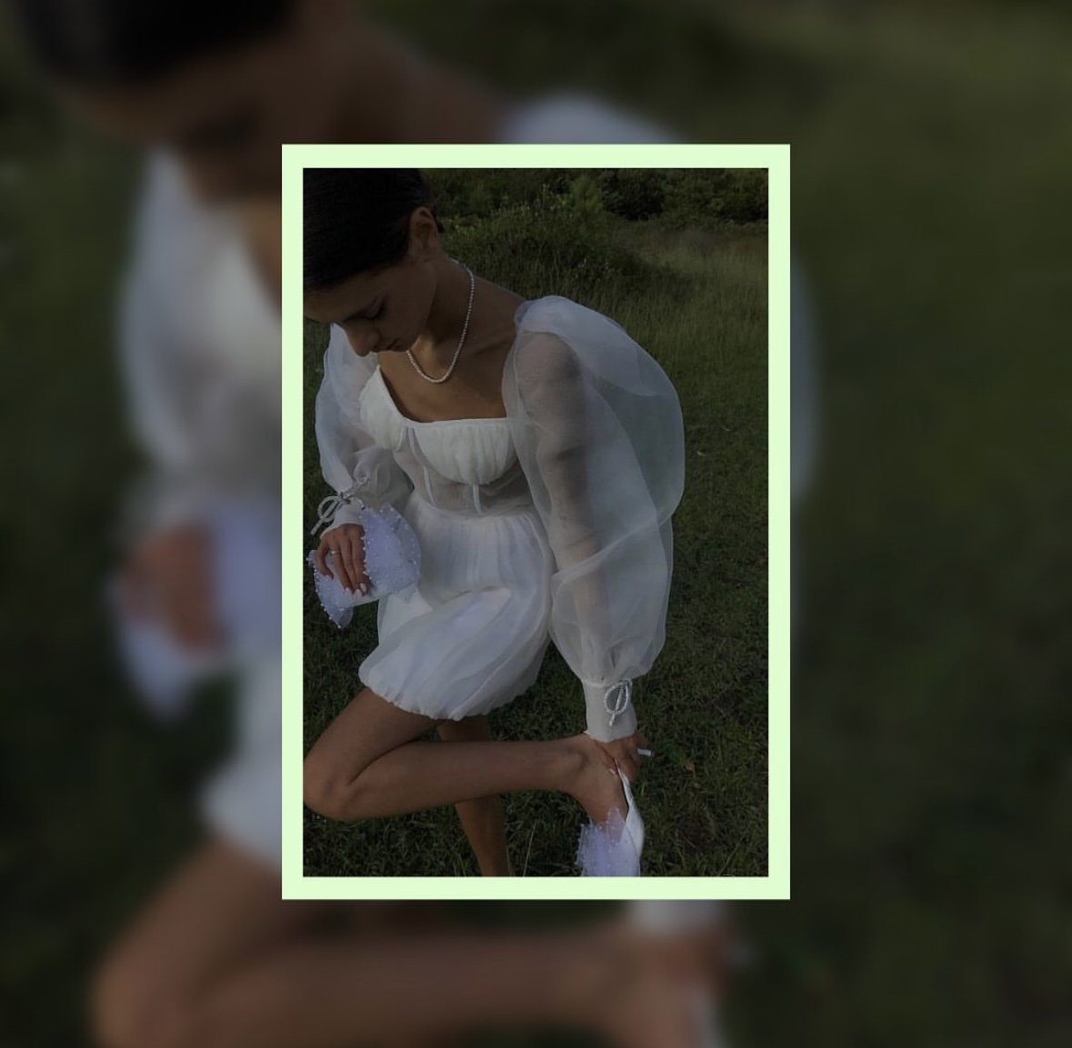 A woman in a white dress with puffy sleeves, standing outdoors on grass, looking down and adjusting her shoe, holding a small, beaded clutch bag.