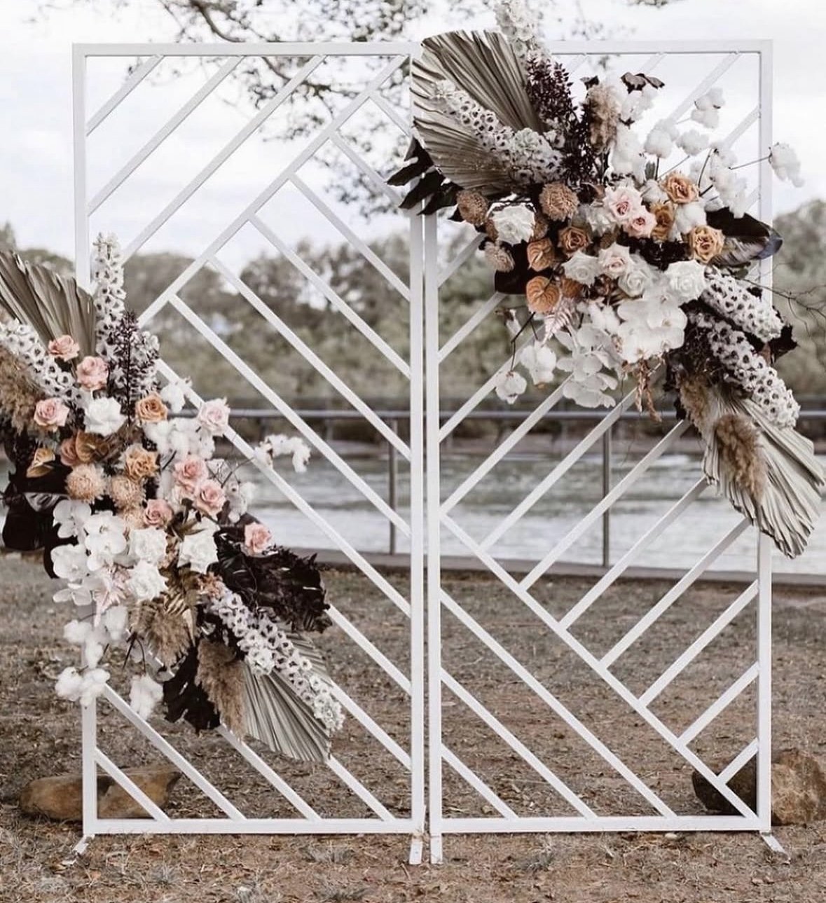 A geometric white metal structure decorated with floral arrangements, set outdoors near a body of water and trees.