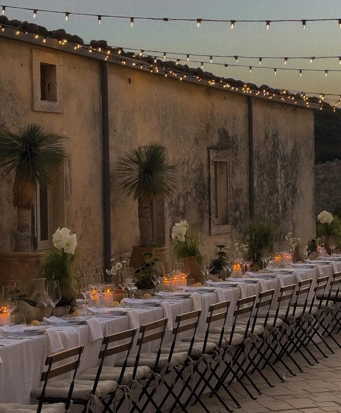 Outdoor dinner setup at dusk with a long table draped in white tablecloths, decorated with flower arrangements, candles, and glassware, against an old stone wall with string lights overhead.