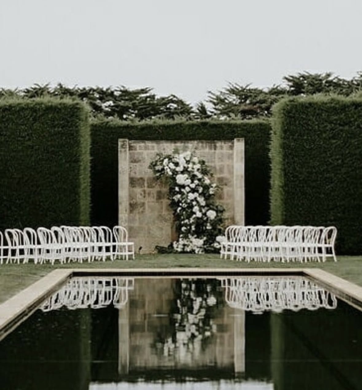 Wedding altar with white chairs arranged on either side, floral arrangement on a stone wall, and reflecting pool in the foreground, set in a garden with tall green hedges.