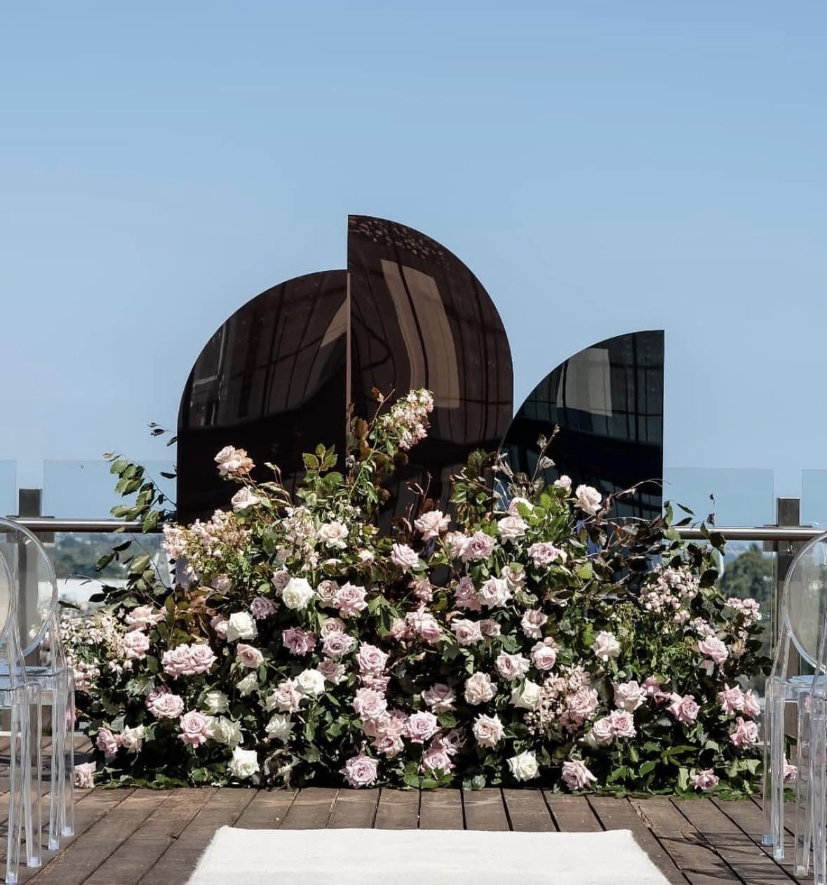 Decorative floral arrangement with pink and white roses in front of a modern black sculpture on a rooftop deck. Clear chairs are visible on each side, with a white rug and a glass railing against a blue sky.