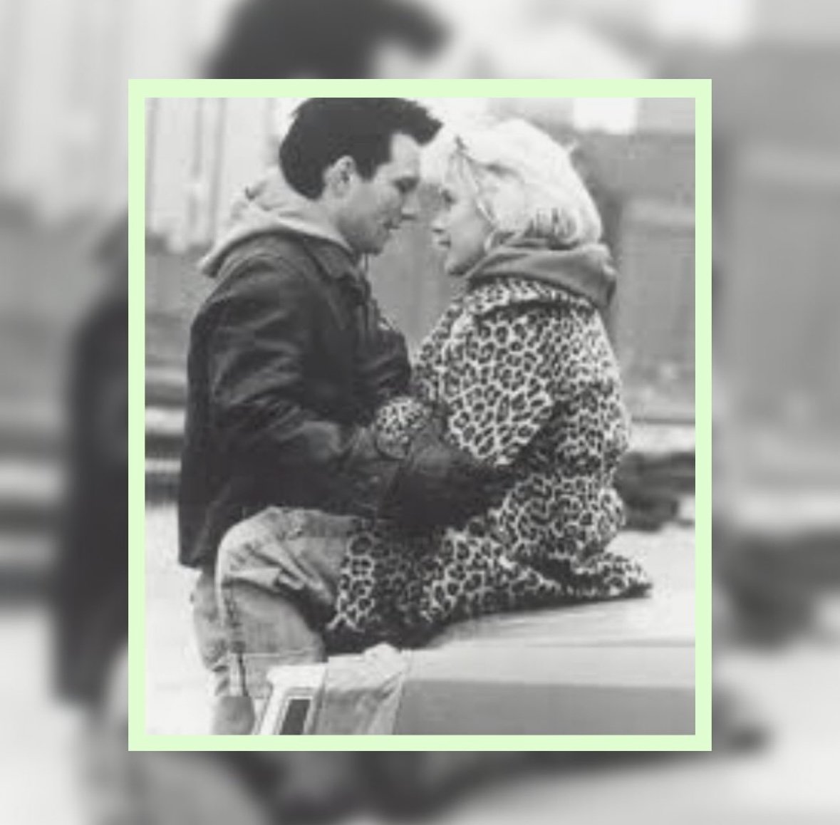 A black and white photo of a young man and young woman sitting closely, facing each other, with foreheads touching. The woman is wearing a leopard print coat and the man is wearing a leather jacket and hoodie. They are outdoors with a blurred backgro