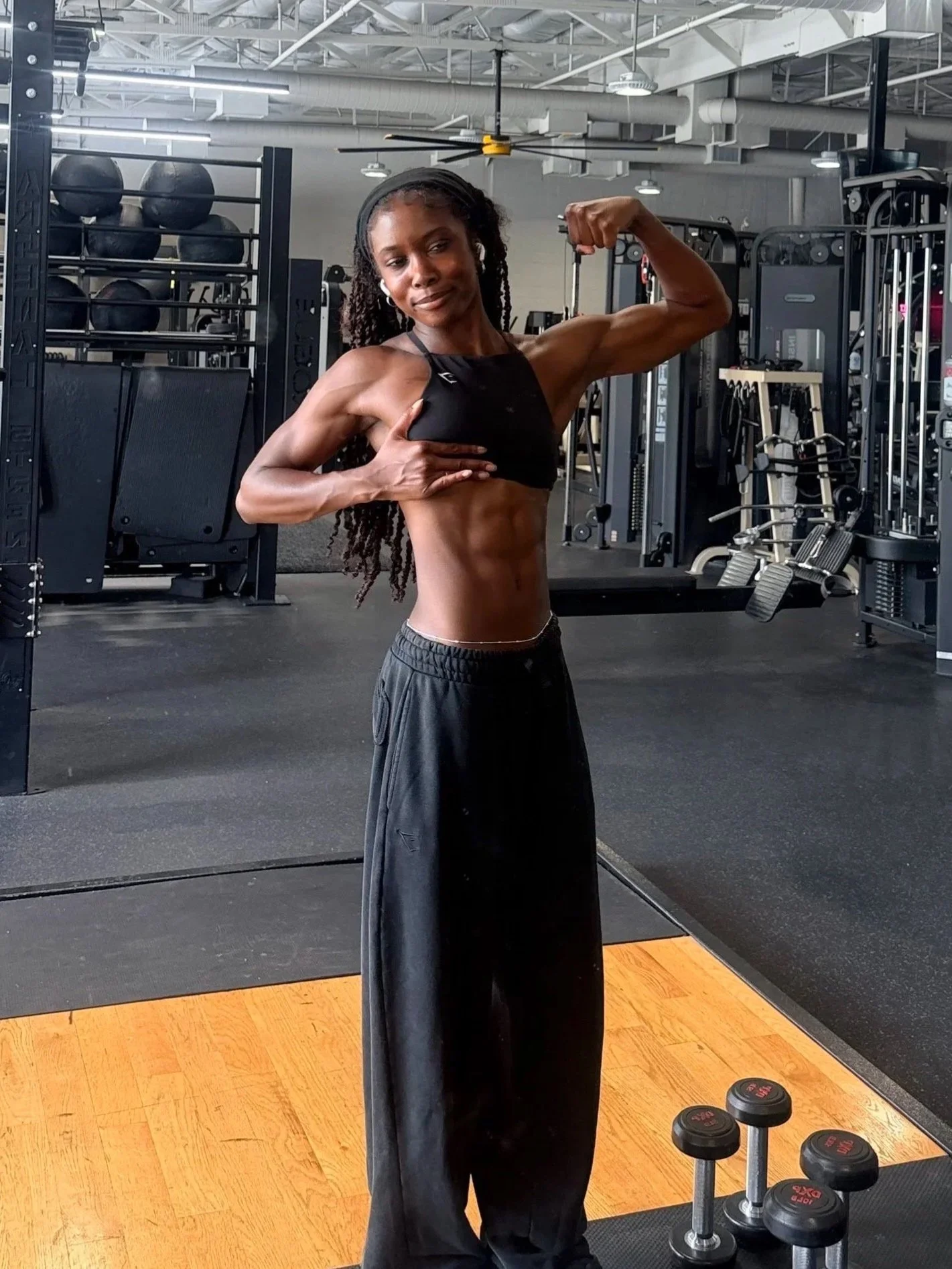 A woman with long braided hair and dark athletic clothing showing off her toned arm muscles in a gym. She is standing in front of gym equipment including dumbbells and a weight machine, with a serious expression.