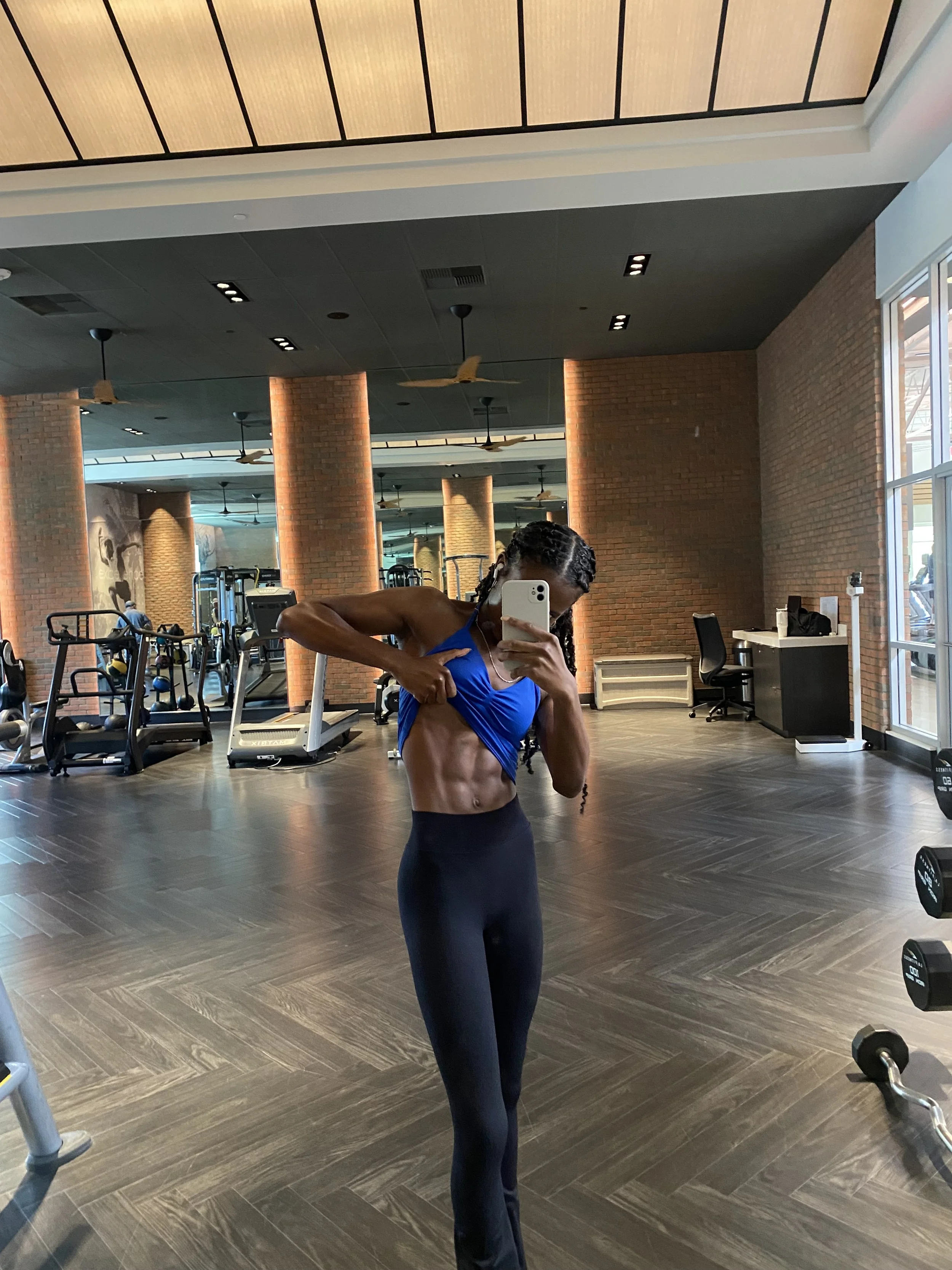 A woman taking a mirror selfie in a gym, wearing a blue sports top and black leggings, with workout equipment and brick walls in the background.