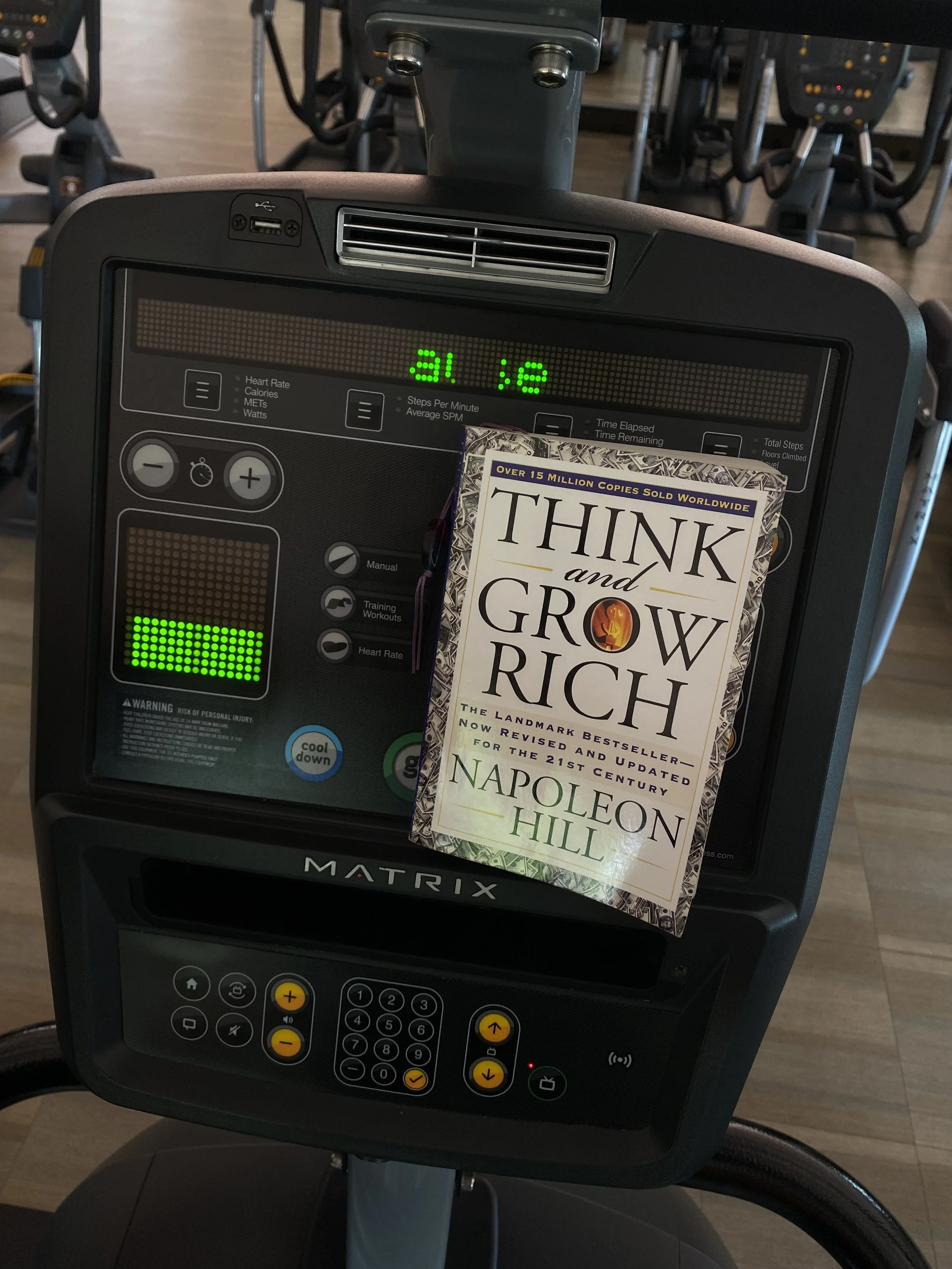 Indoor gym setting with exercise bikes, a digital display on one bike indicating steps, and a book titled 'Think and Grow Rich' by Napoleon Hill placed on the console of a bike.