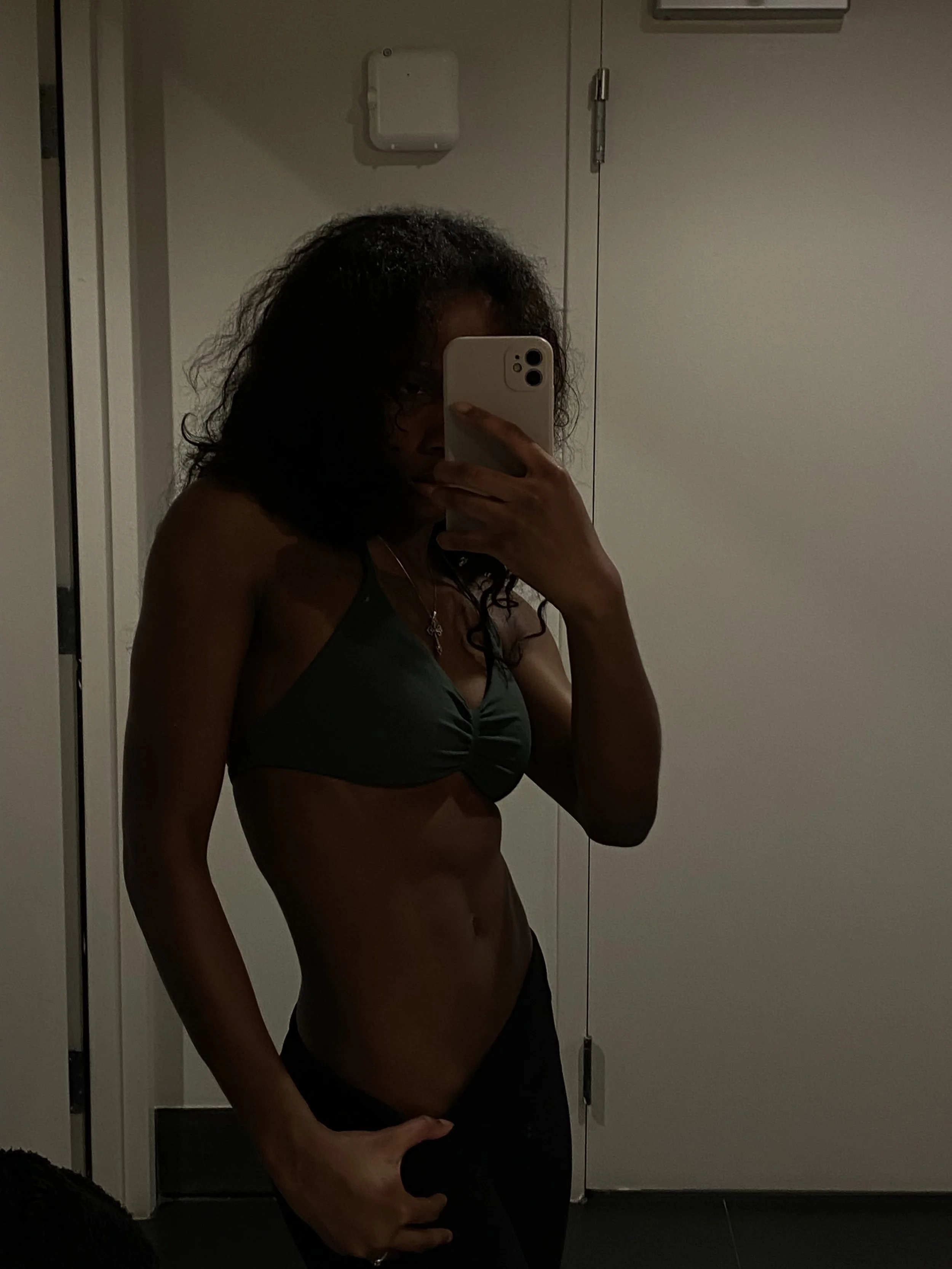 A person taking a mirror selfie in a dimly lit room, wearing a sports bra and underwear, with dark, curly hair partially obscuring their face.