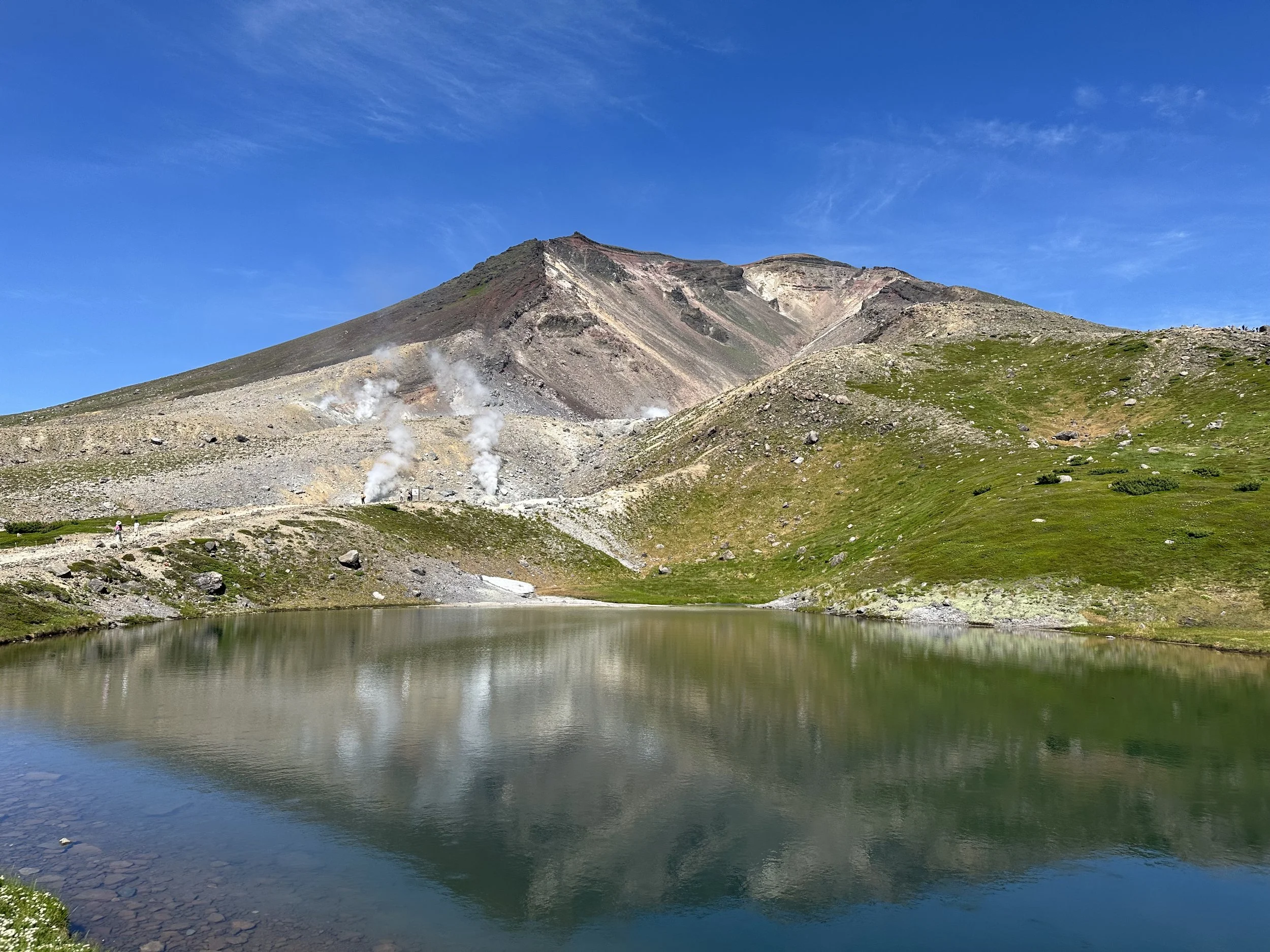 A mountain landscape with a lake in the foreground, green hills on either side, a steep mountain behind, and steam rising from fumaroles on the mountain slope, under a bright blue sky.