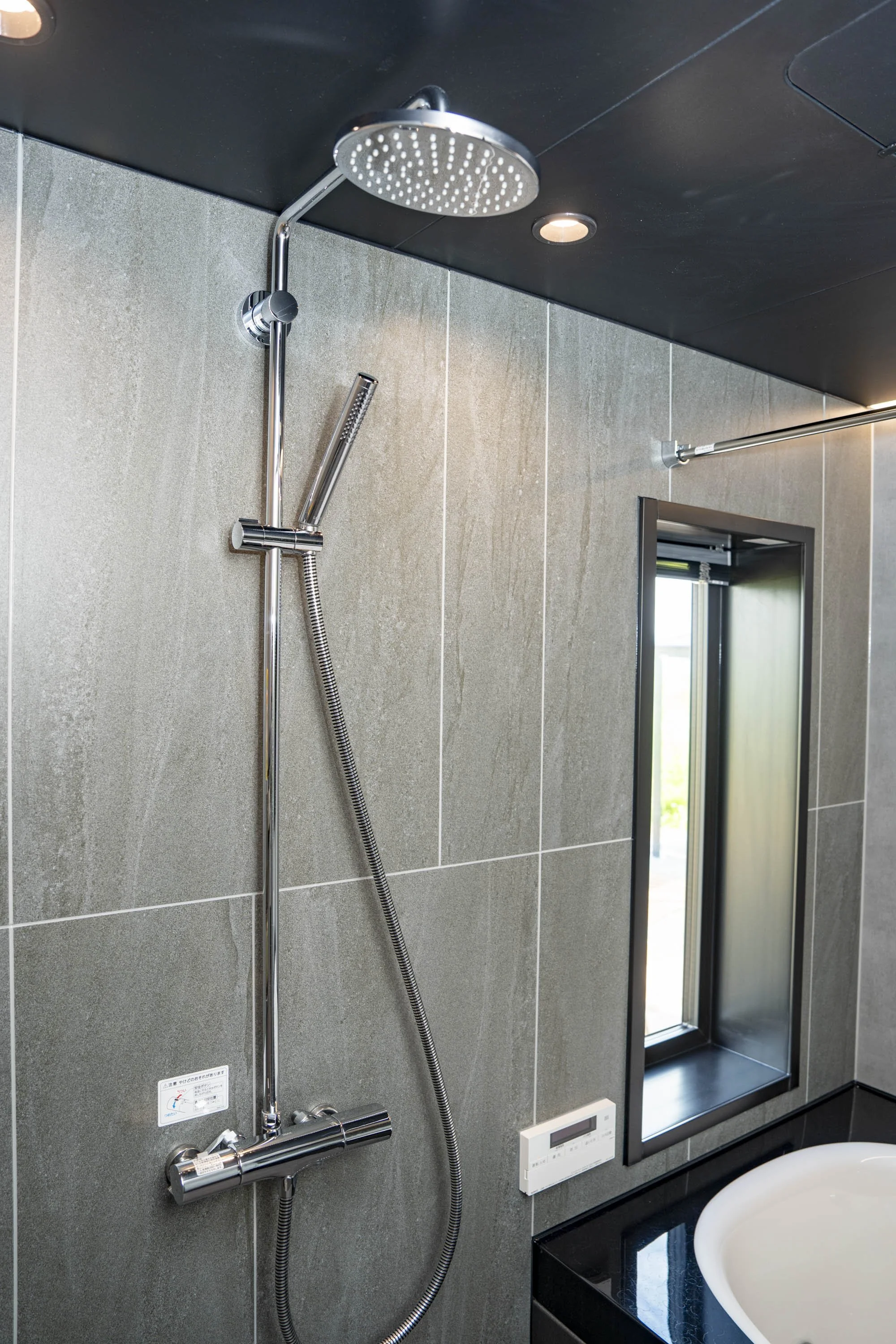 Modern bathroom with a wall-mounted showerhead, handheld shower, a small window, and a control panel on a gray tiled wall.