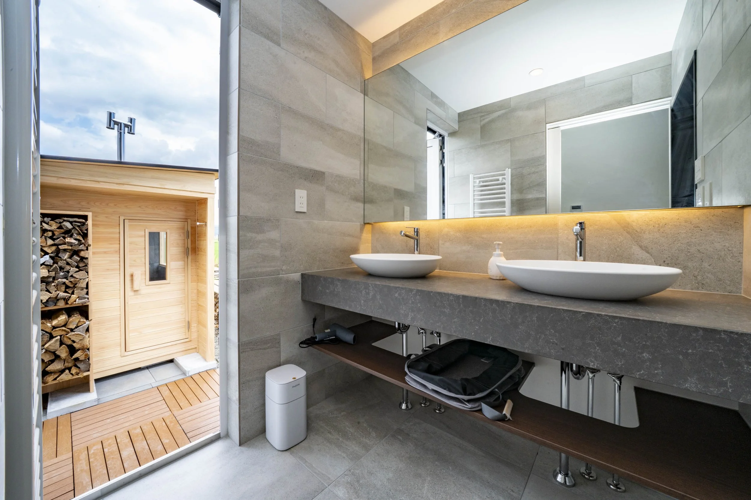 Modern bathroom with double vessel sinks, large mirror, and outdoor sauna with wood storage outside.