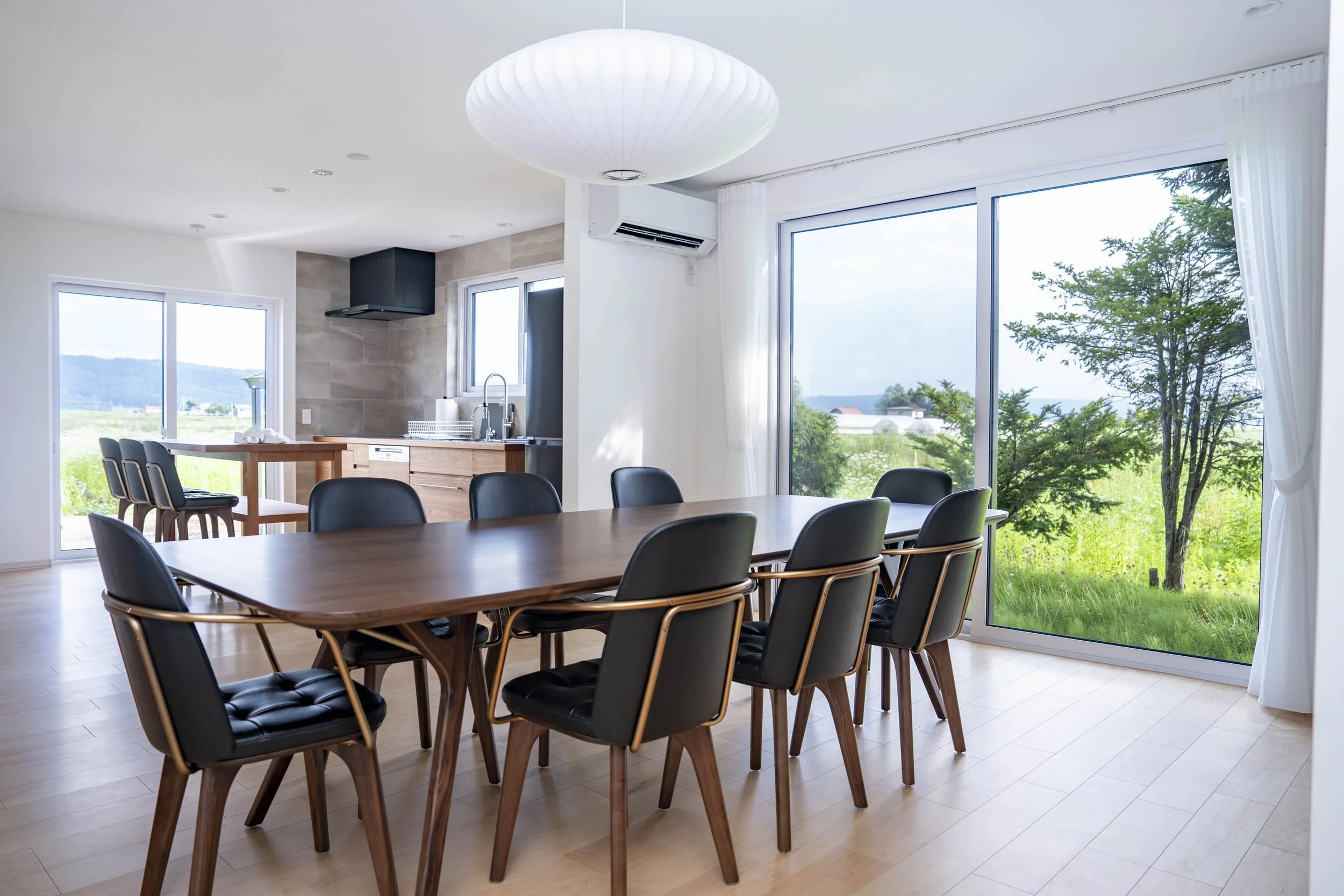 Modern dining room with a wooden table and six black and wood chairs, large glass sliding doors with view of green landscape, in a bright, minimalist kitchen.