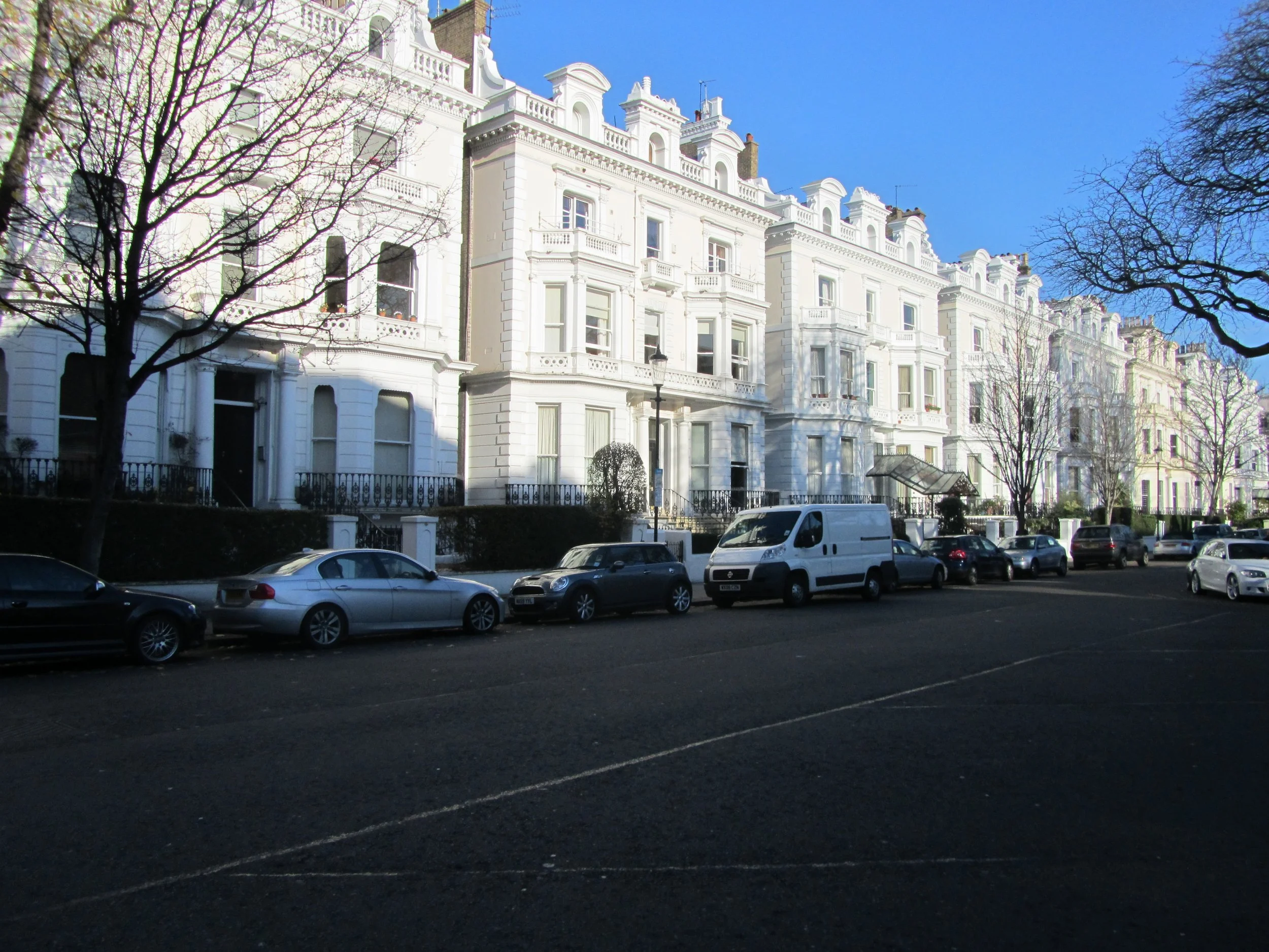 Street view with white Victorian-style row houses, parked cars, and leafless trees under a clear blue sky.