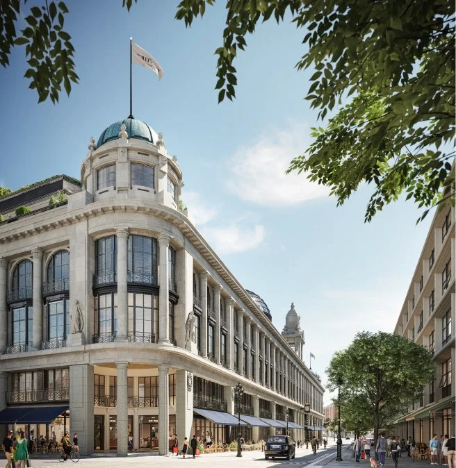 A historic building with classical architecture, featuring a rounded corner, large windows, and a rooftop flag, situated on a lively city street with trees and pedestrians.