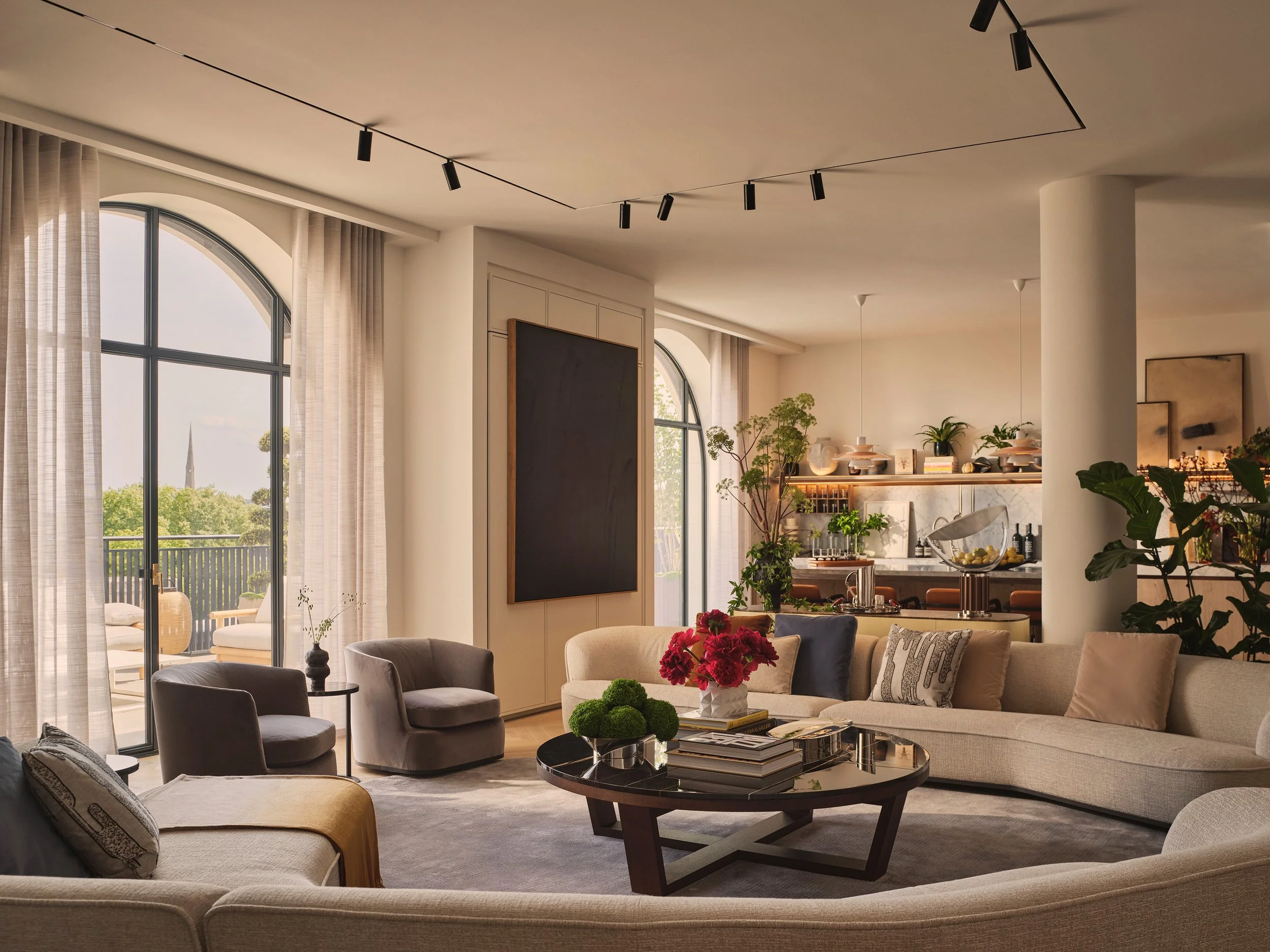 Spacious living room with beige couches, armchairs, a round coffee table with books and flowers, large glass doors with curtains leading to an outdoor balcony, and a kitchen area with plants and decorations in the background.