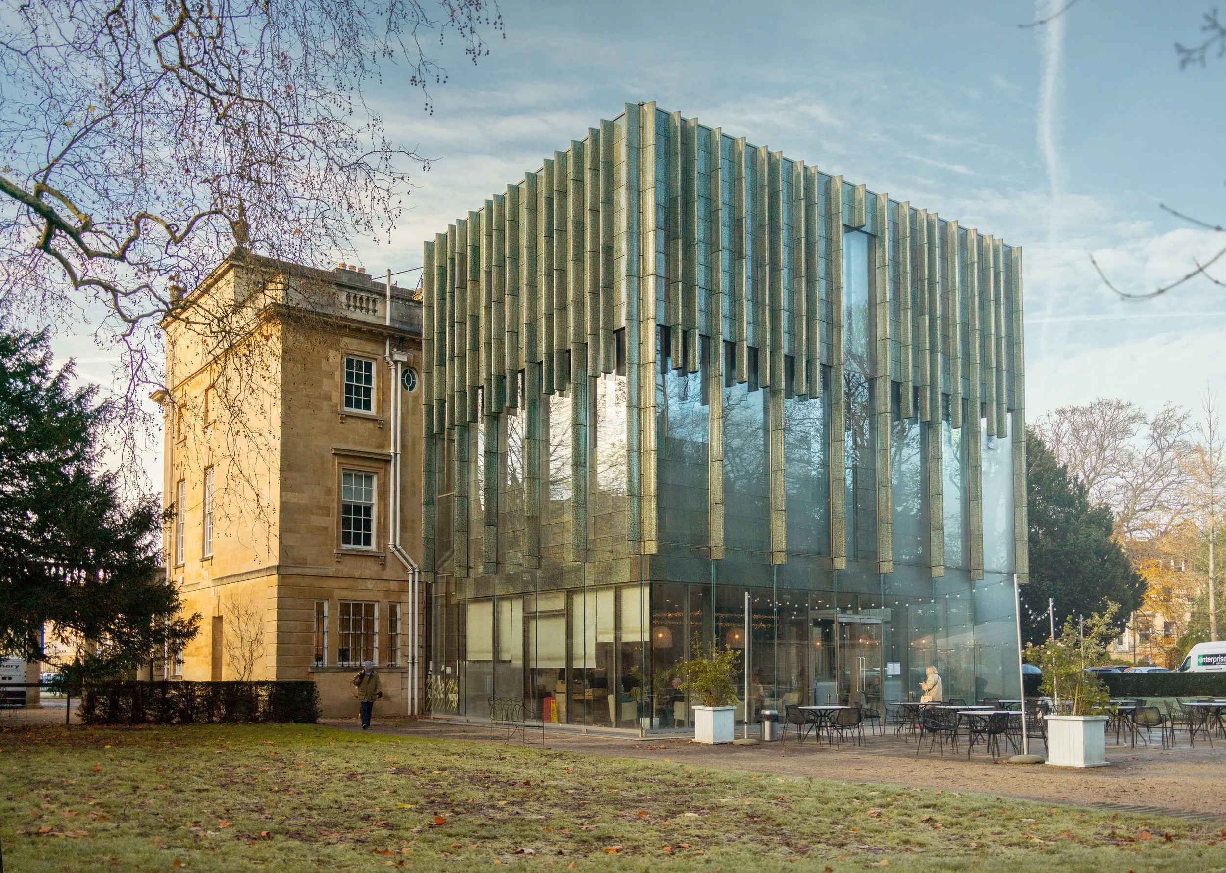 Modern glass building with vertical metal accents next to an older stone building in a park with trees and outdoor seating.
