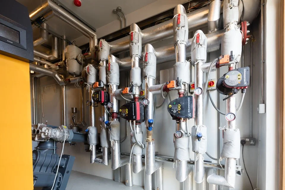 Complex industrial piping system with insulated pipes, gauges, and control modules in a mechanical room.