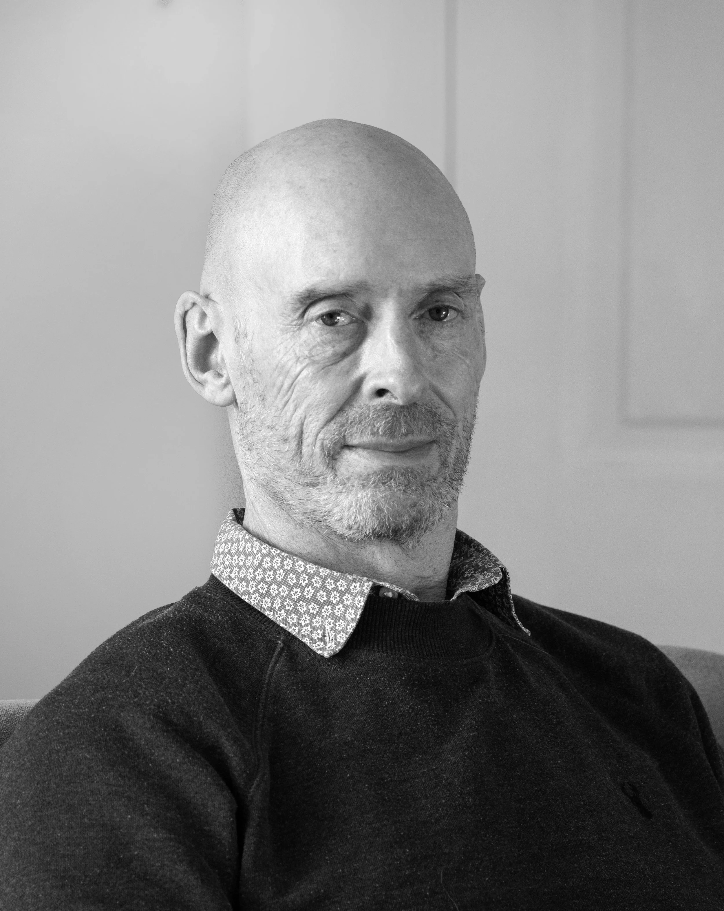 Black and white photo of a bald man with a beard, wearing a patterned collared shirt under a dark sweater, sitting on a couch.